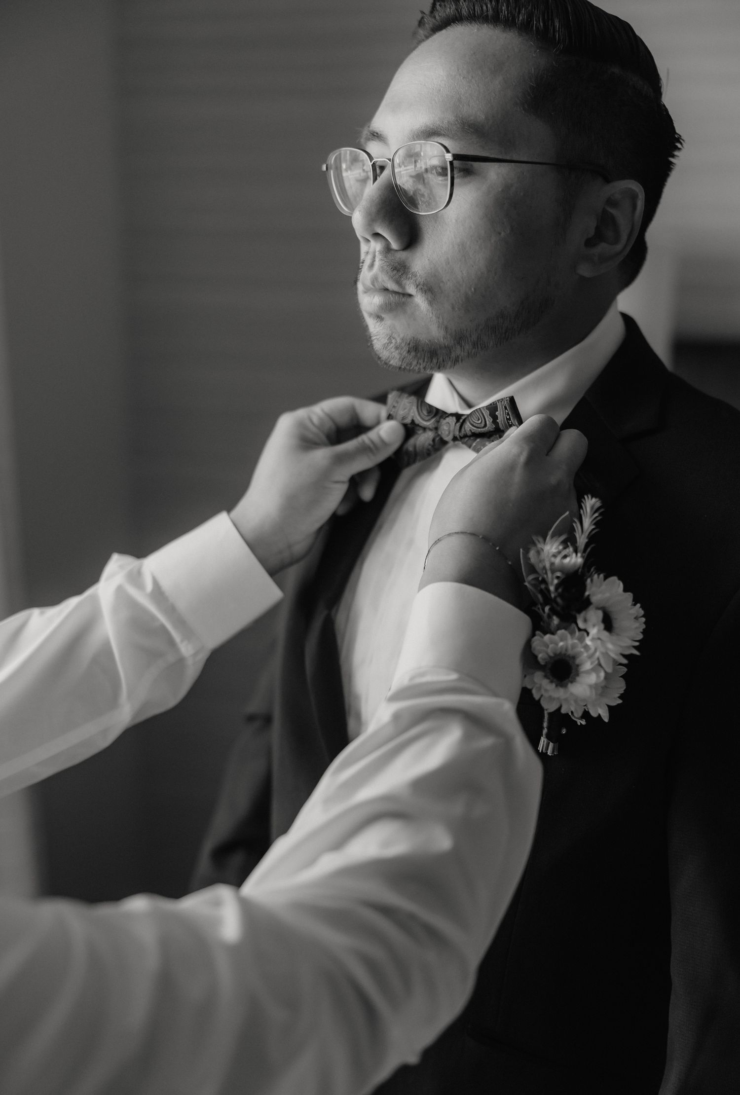 Black and white portrait of someone having their bow tie adjusted while wearing glasses and a boutonniere.