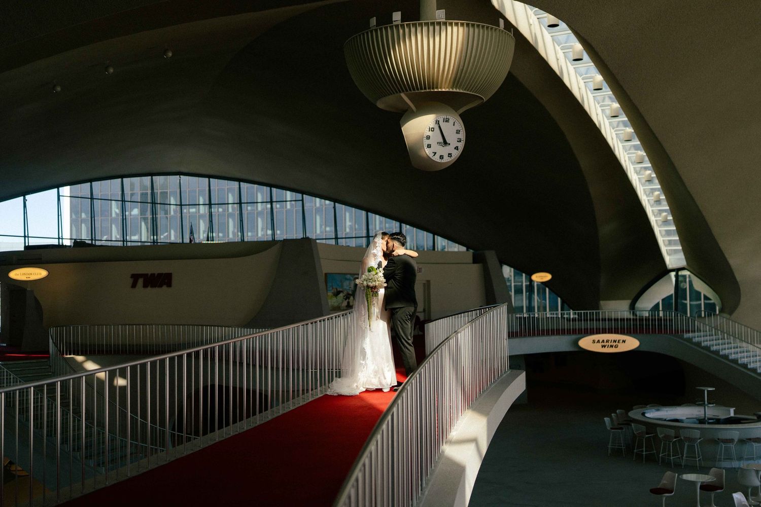 A bride and groom embrace in an elegant pose against a minimalist white architectural corner backdrop.