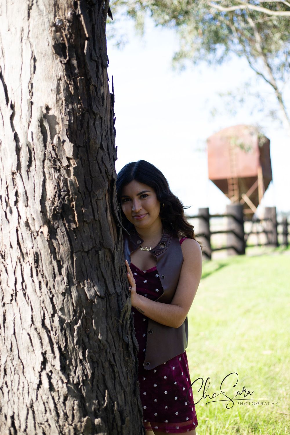 Woman in polka dot dress poses playfully behind tree trunk in sunny outdoor setting.
