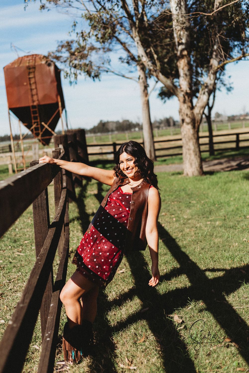Woman in red polka dot dress poses energetically near wooden fence in rural setting.