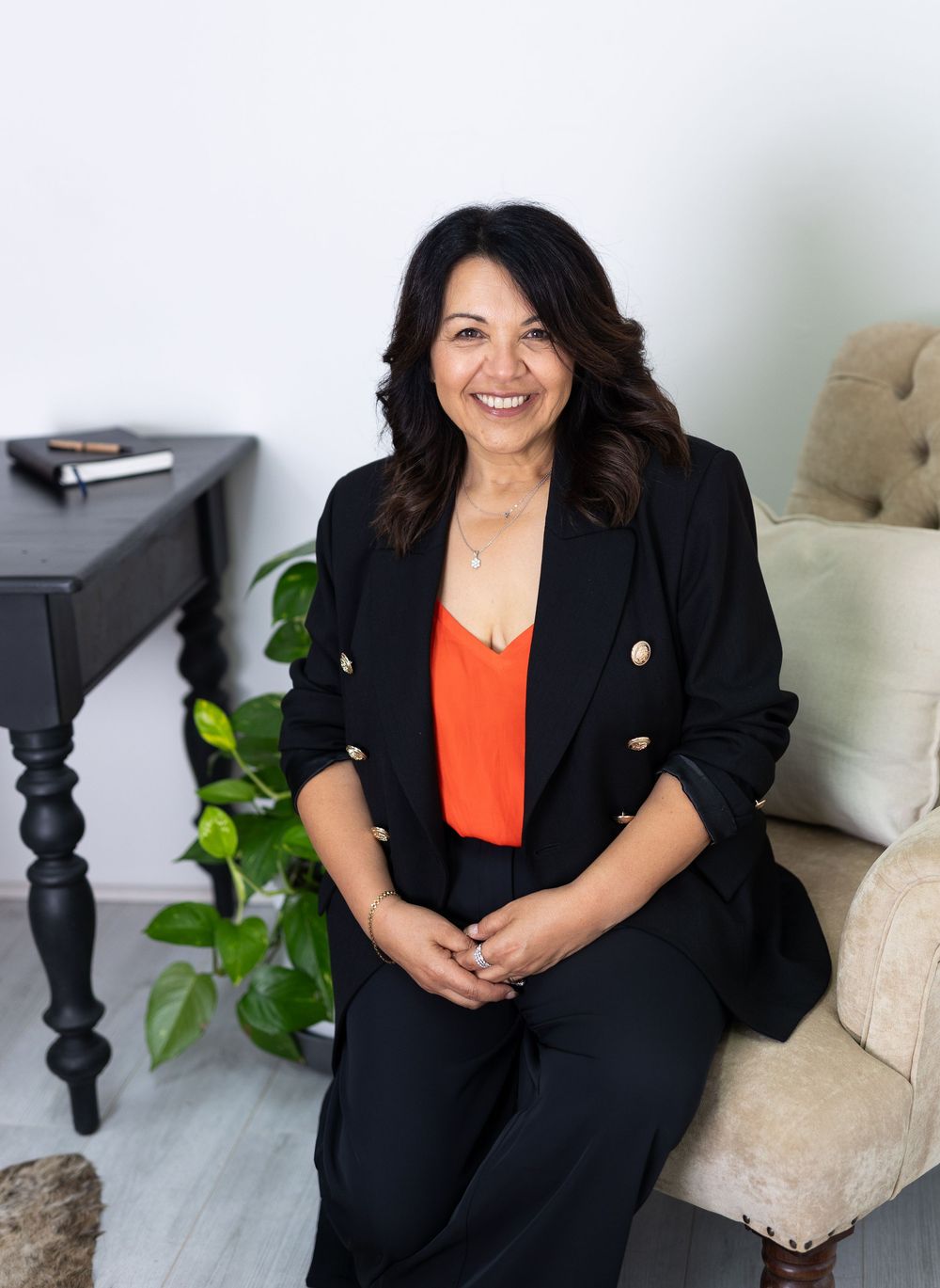 Business professional in black blazer and orange top sitting on couch in modern office setting.