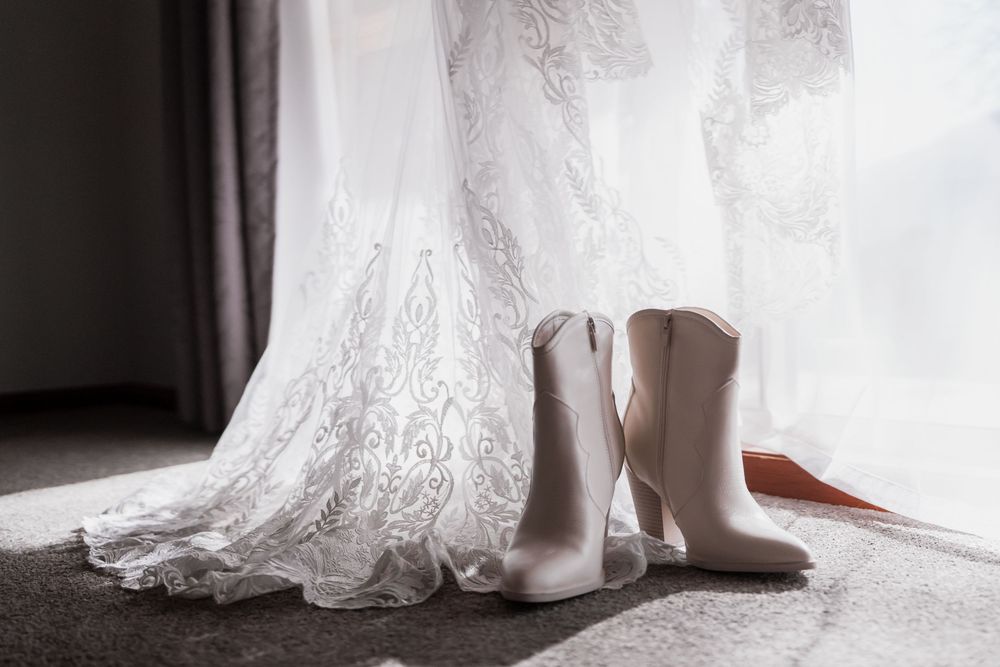 White cowboy boots sit beside flowing lace wedding dress against soft gray background.