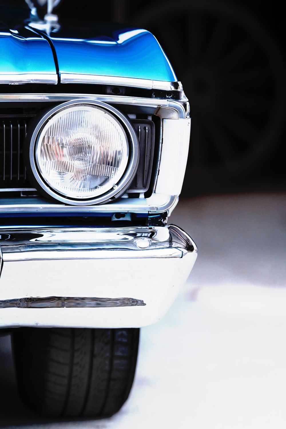 Classic vintage car headlight and front grille detail with chrome trim and blue paint in dramatic lighting.
