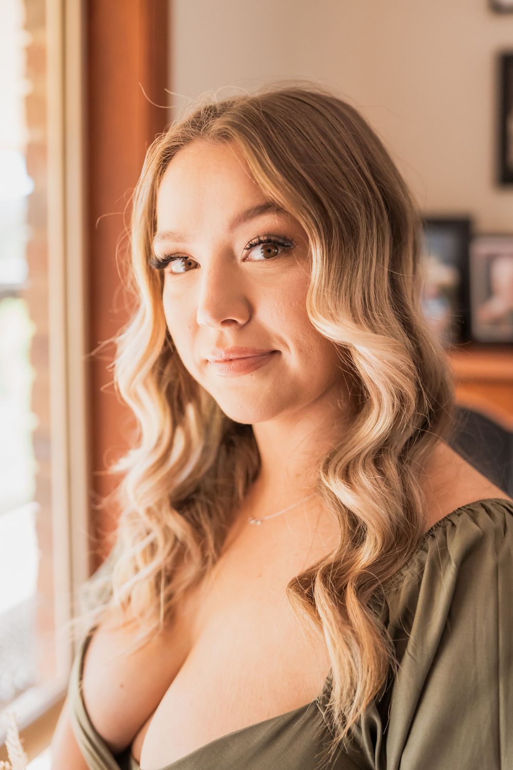 Portrait of a person with wavy blonde hair and olive top by window with soft natural lighting.
