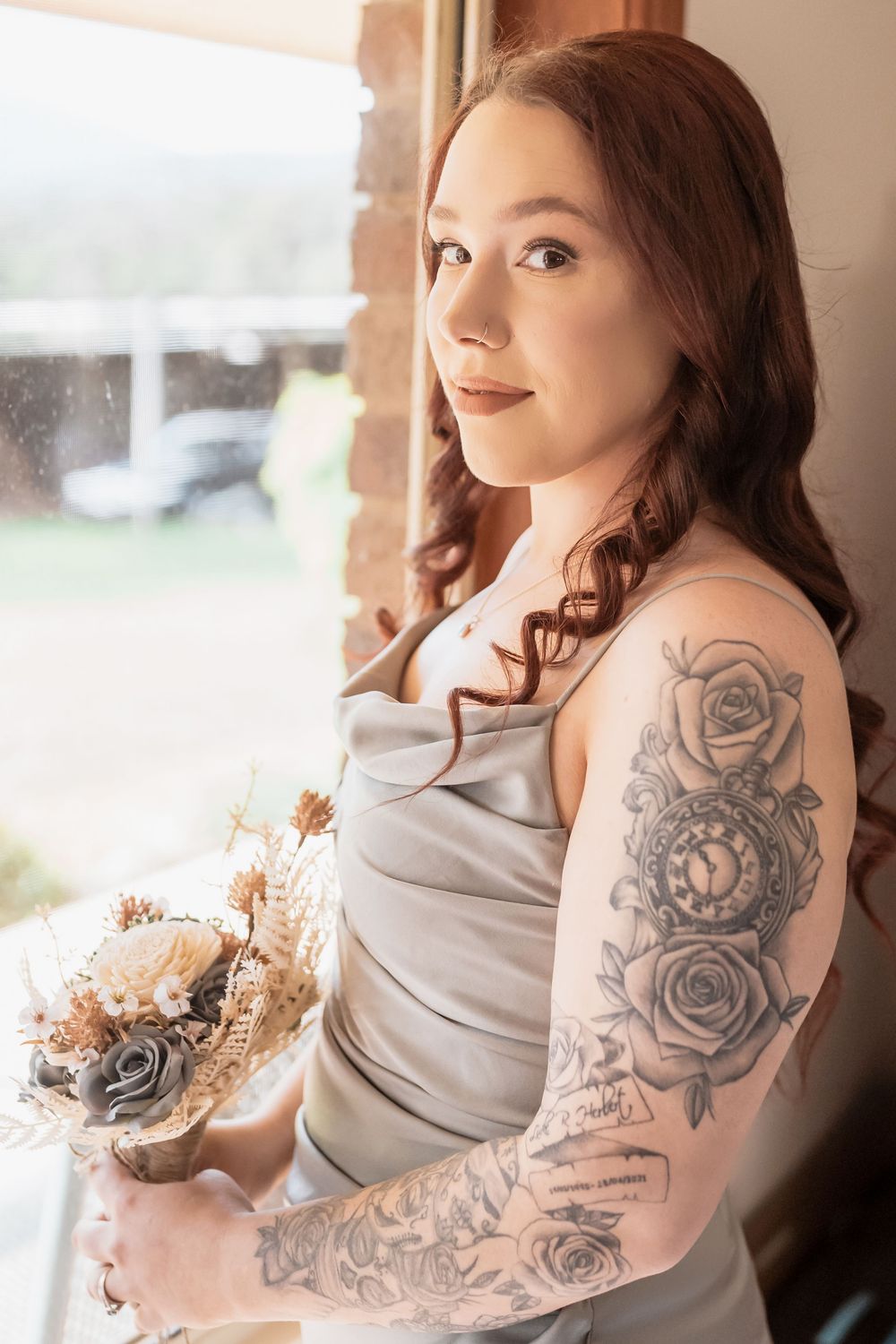 A bride in a light dress holds a muted bouquet by a window, showing beautiful floral tattoo sleeve artwork on her arm.