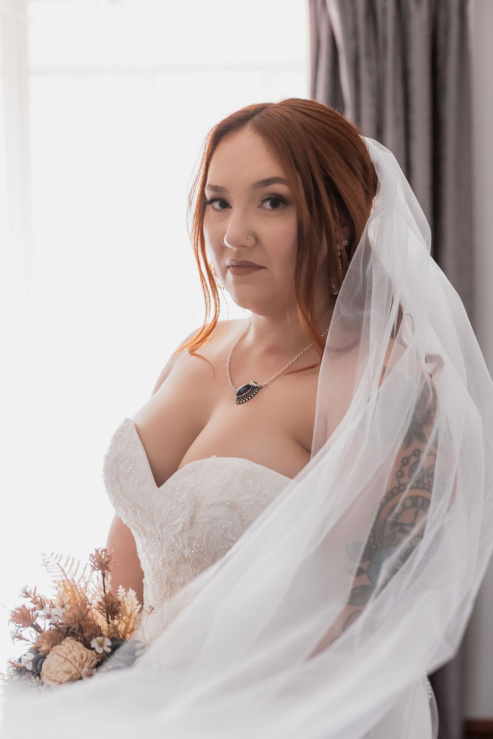 A bride in a white strapless wedding dress poses with a flowing veil and dried flower bouquet indoors.