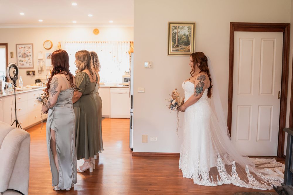 A bride wearing a white lace wedding gown with a flowing train stands in a sunlit room with bridesmaids nearby.