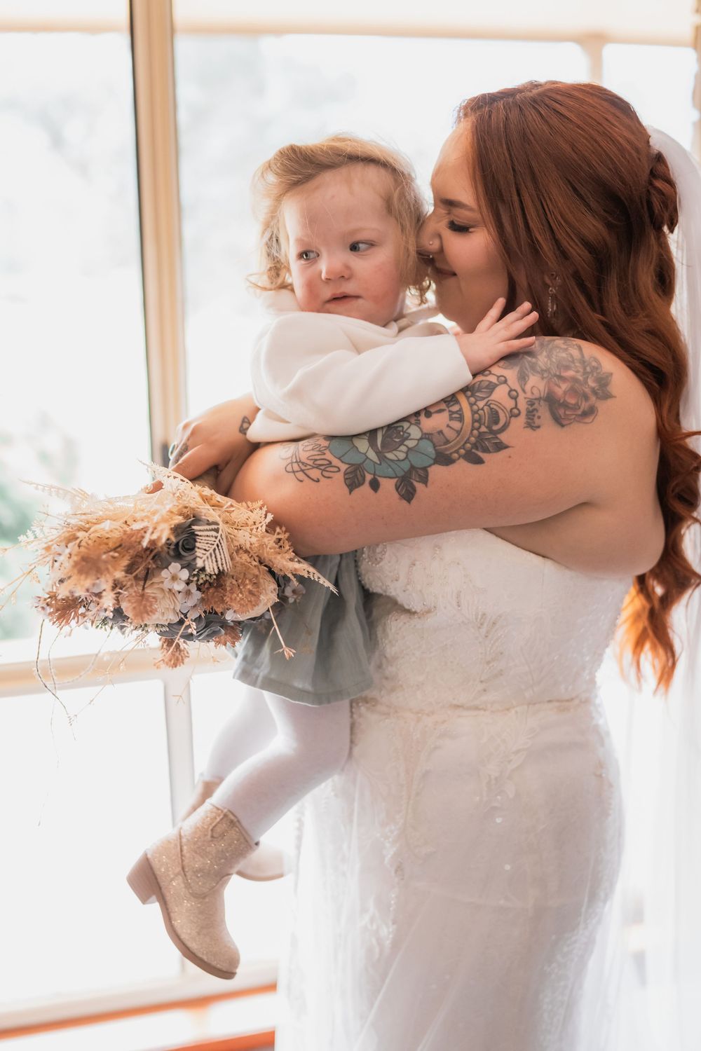 Person in a white lace dress holds and embraces small child near a bright window with visible floral-style tattoo.