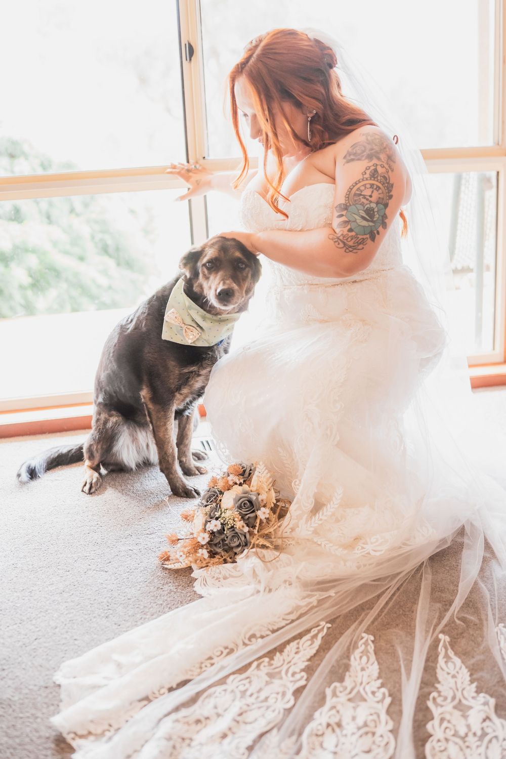 A bride in a lace wedding dress sits with her dog wearing a bandana by a sunlit window.