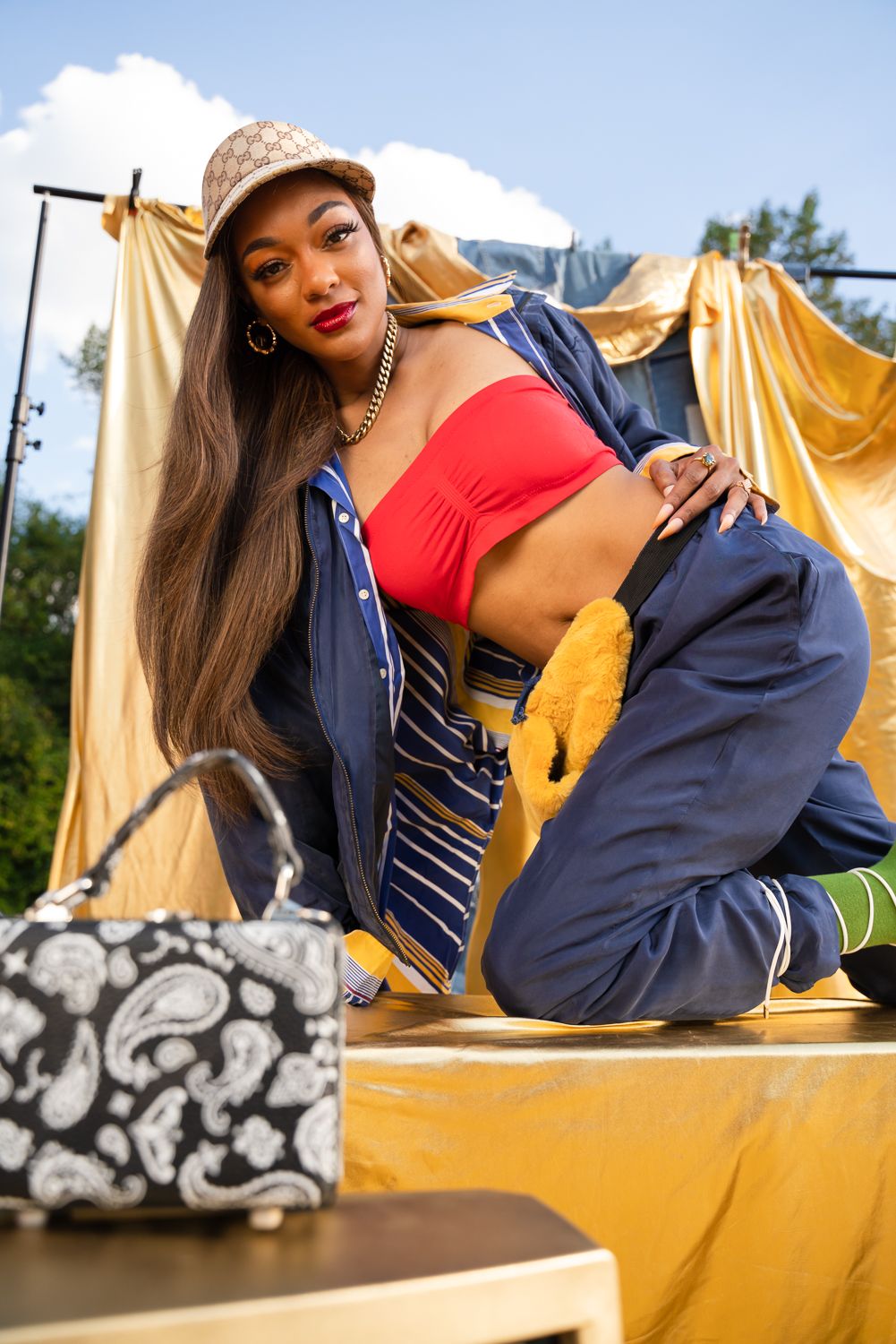 Model in navy and red outfit poses on wooden platform against gold backdrop outdoors.