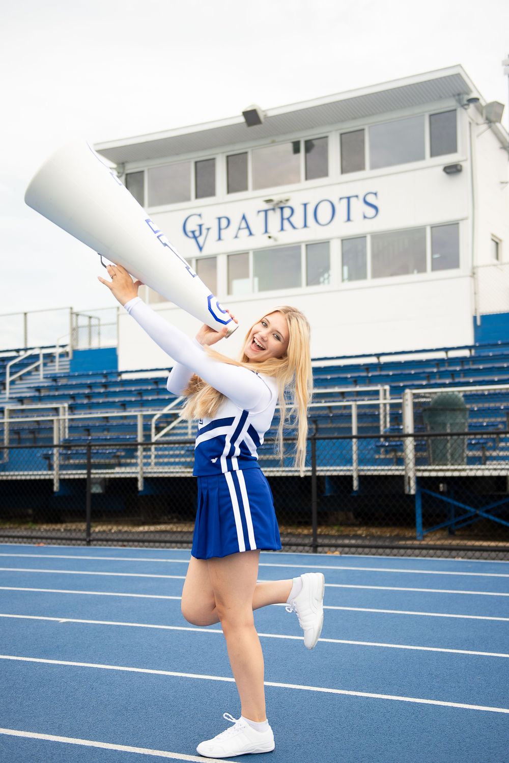 Cheerleader in blue and white uniform performs stunts on an outdoor track with "Patriots" text visible in background.