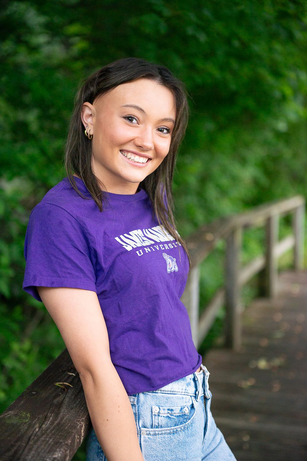 A smiling person wearing a purple collegiate t-shirt and jeans leans against a wooden railing in an outdoor setting with greenery.