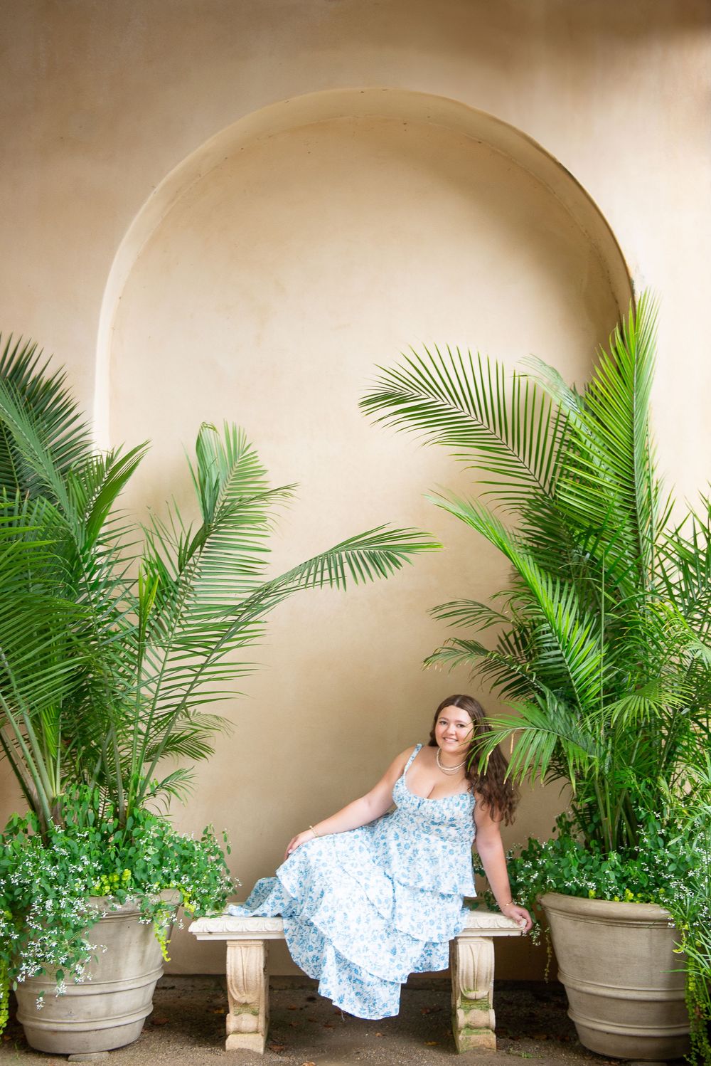 A figure in a flowing blue dress poses dynamically against a curved wall with tropical palm plants in gray planters.