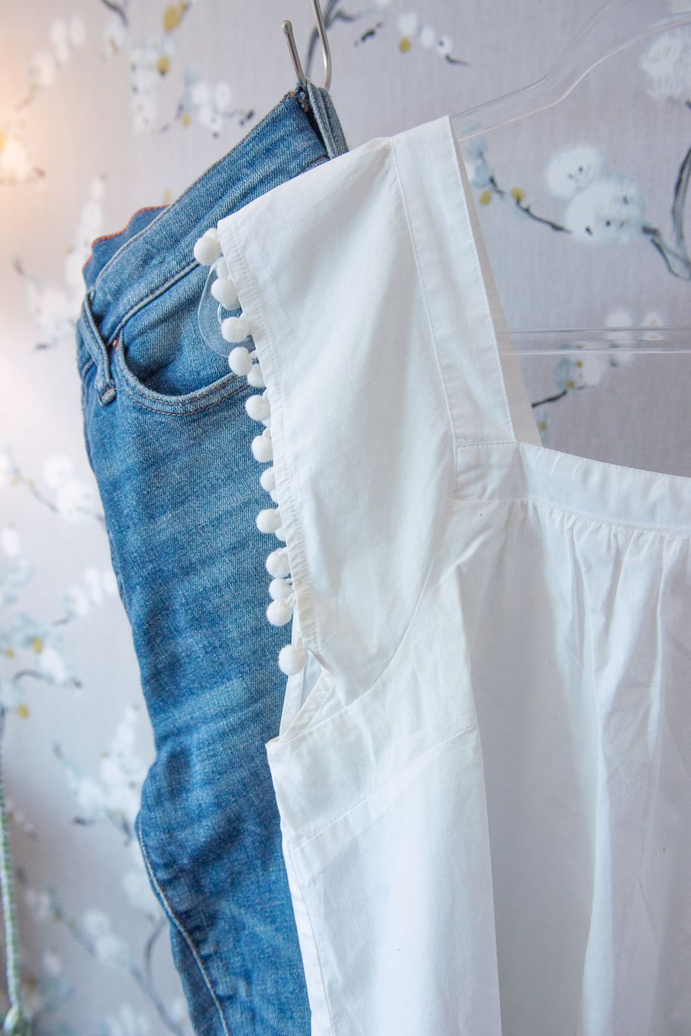 Blue denim jeans and white cotton blouse with pom pom trim hanging against a light background with string lights.