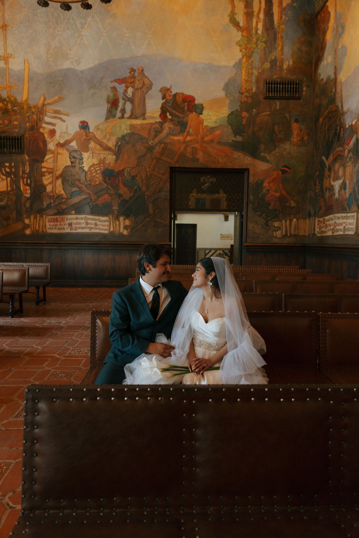 A romantic scene shows a bride and groom sitting together on an ornate bench with decorative murals in the background.