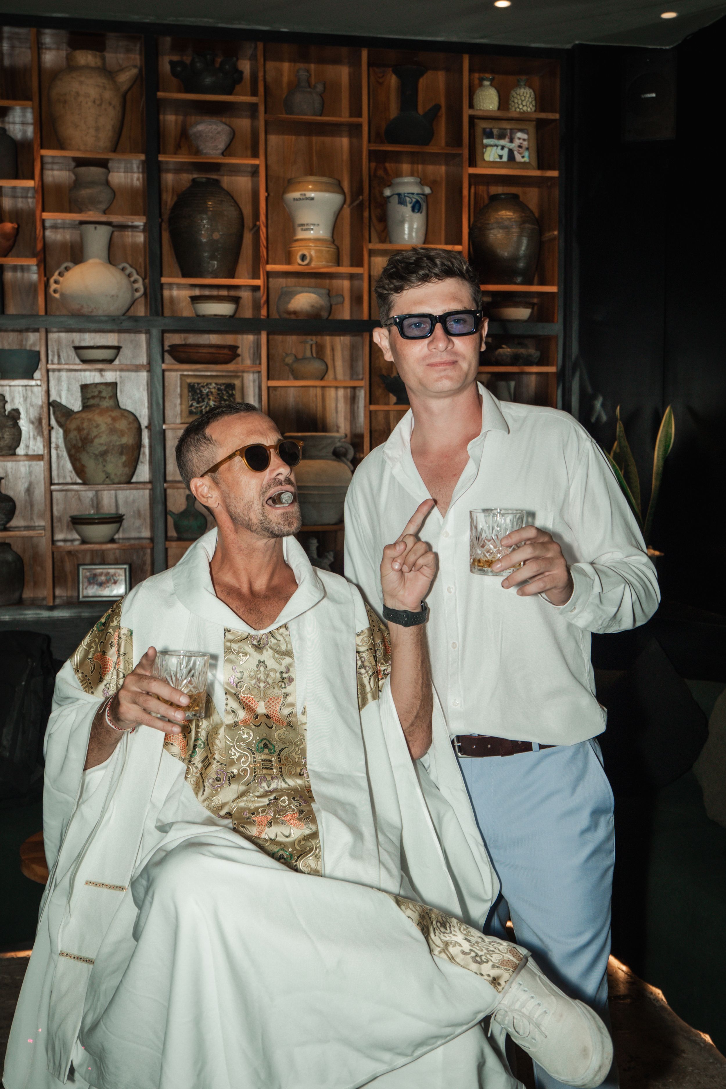 Two people enjoying drinks in a casual setting with vintage pottery and vases displayed on wooden shelves in the background.