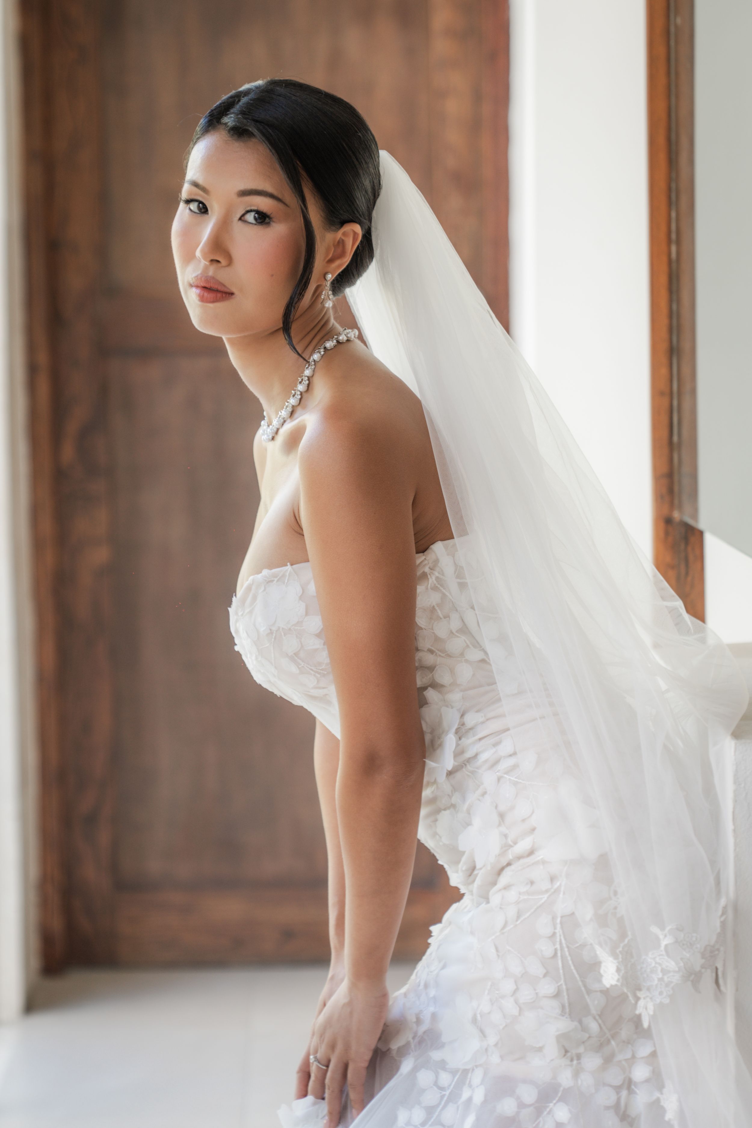 Bride in stunning white lace wedding dress and flowing veil poses elegantly against wooden door in natural light.