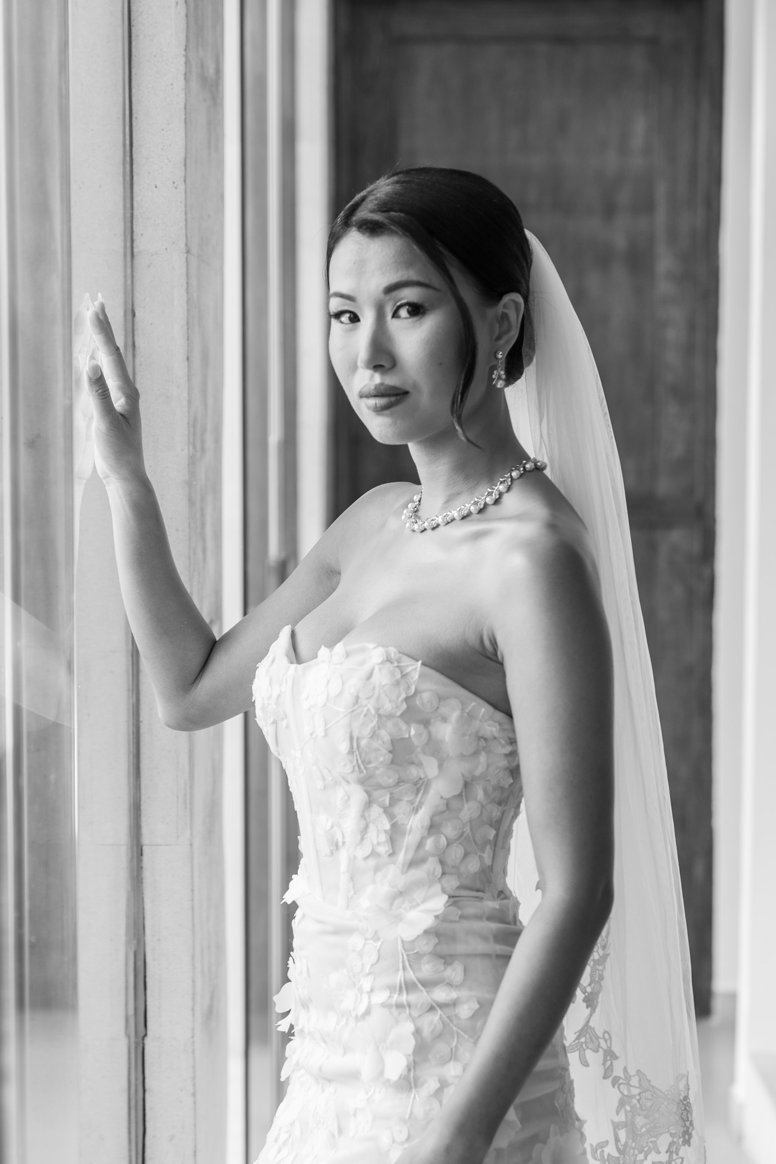 Elegant black and white bridal portrait showing bride in strapless lace gown and veil standing by window.