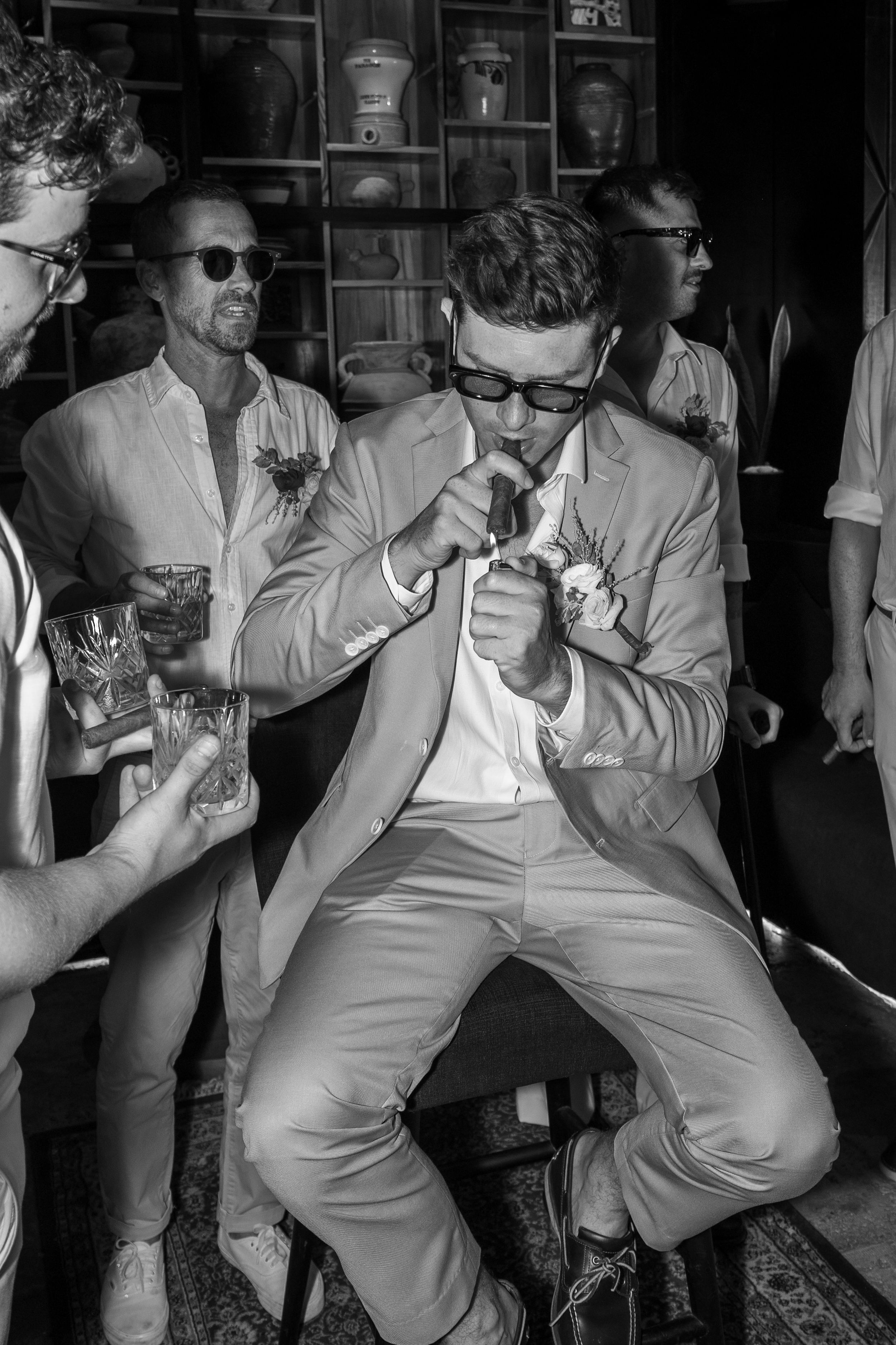Stylish wedding party in sunglasses and light colored suits light up cigars at celebration reception.