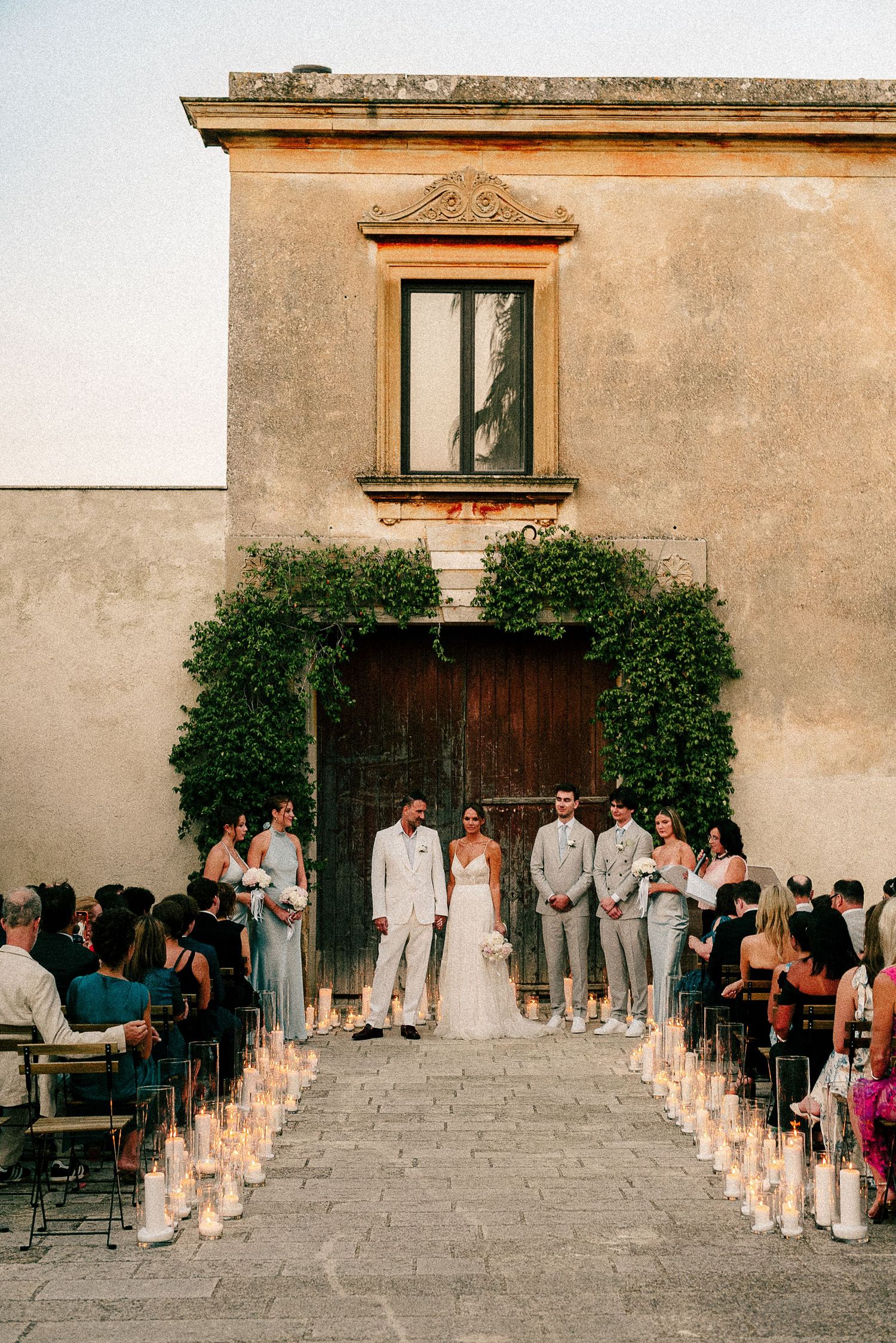 Wedding ceremony takes place at rustic venue with stone walls, wooden door and candlelit aisle lined with guests.