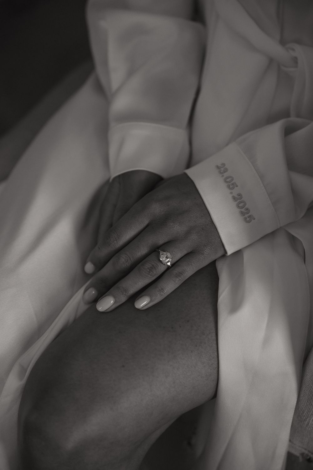 Close-up black and white photo of engagement ring on bride's hand against white fabric.