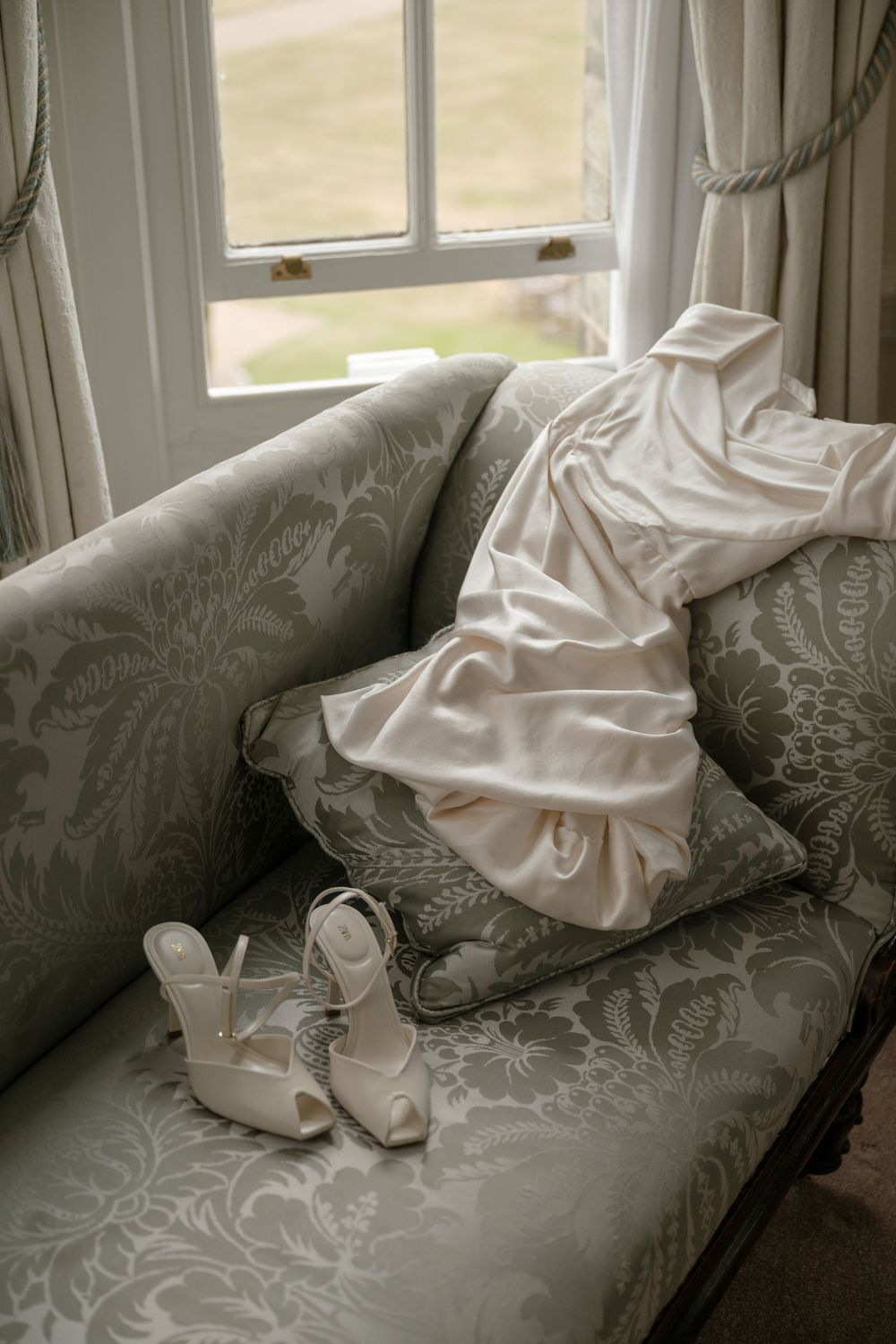 White wedding dress and shoes displayed on gray damask sofa near bright window.