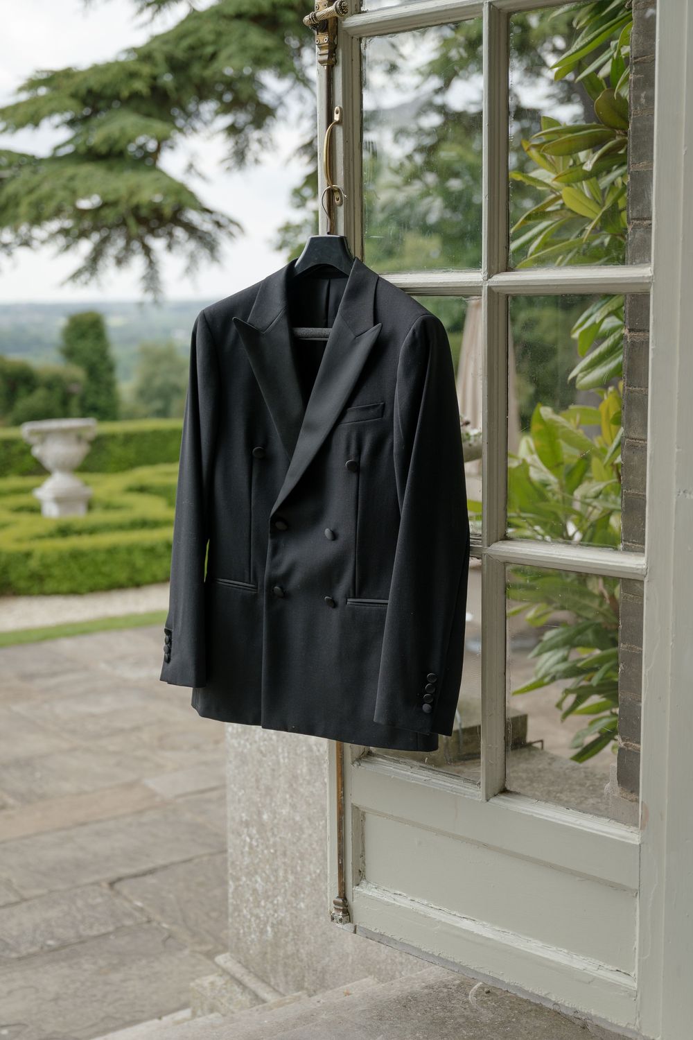 Black tuxedo jacket hangs by window overlooking manicured garden.