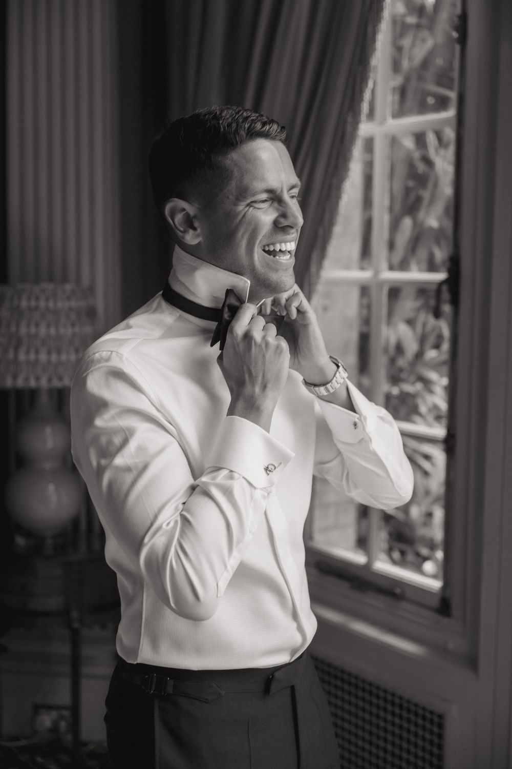 Black and white portrait of groom adjusting bowtie while smiling by window.