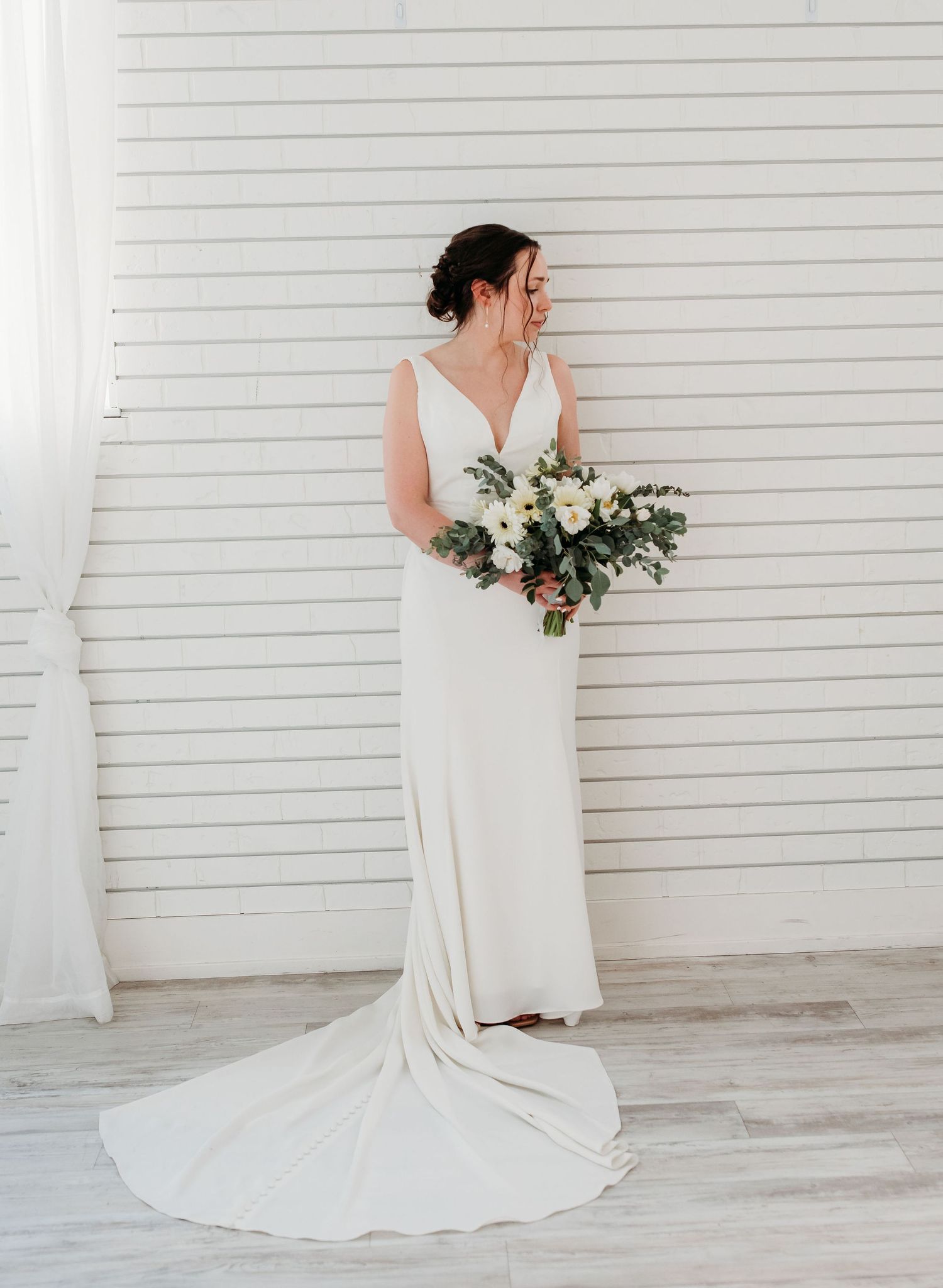 A bride in a flowing white wedding gown with train holds a beautiful bouquet against a white shiplap wall.