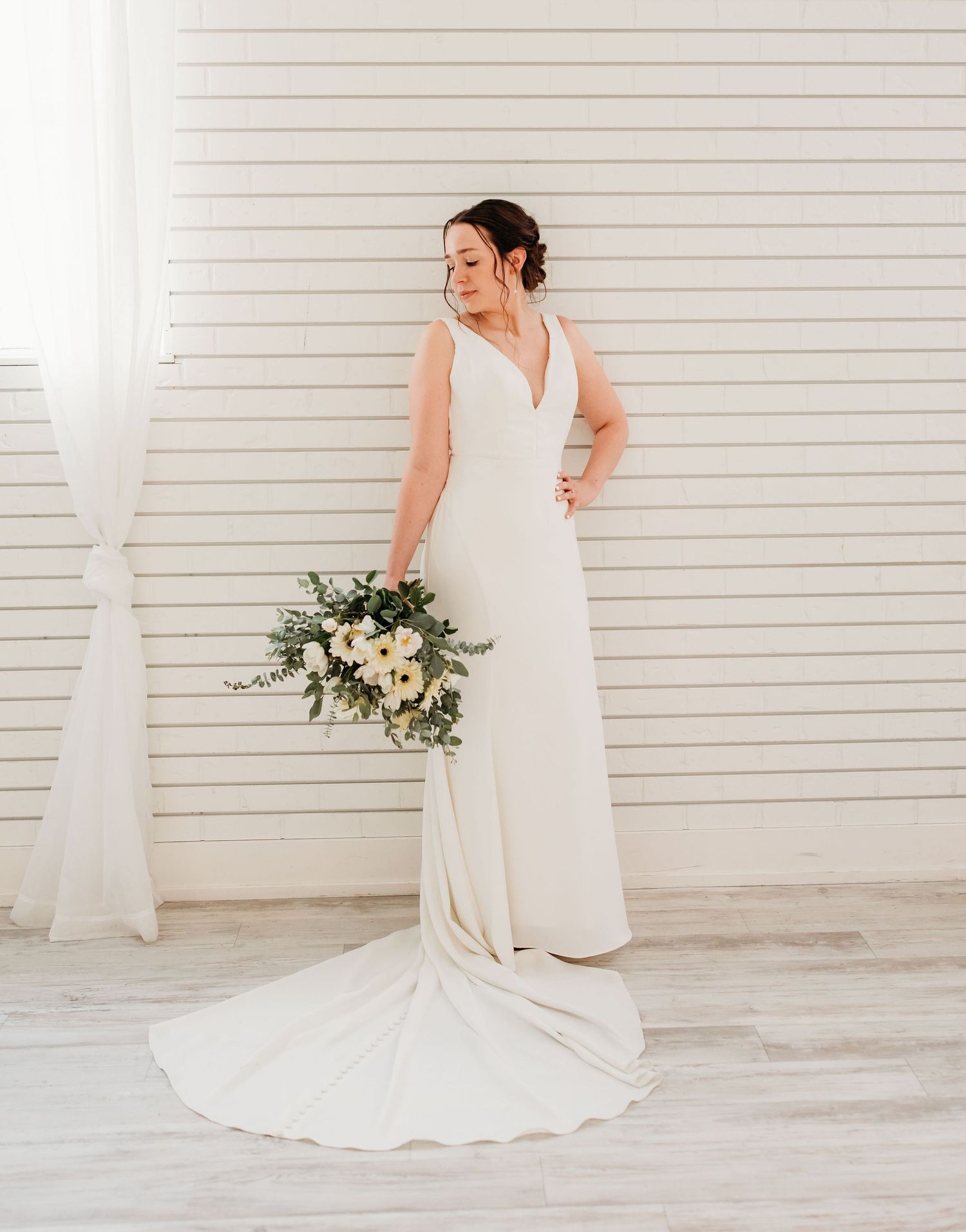 Elegant bride in flowing white V-neck gown with long train holds romantic bouquet against white shiplap wall.