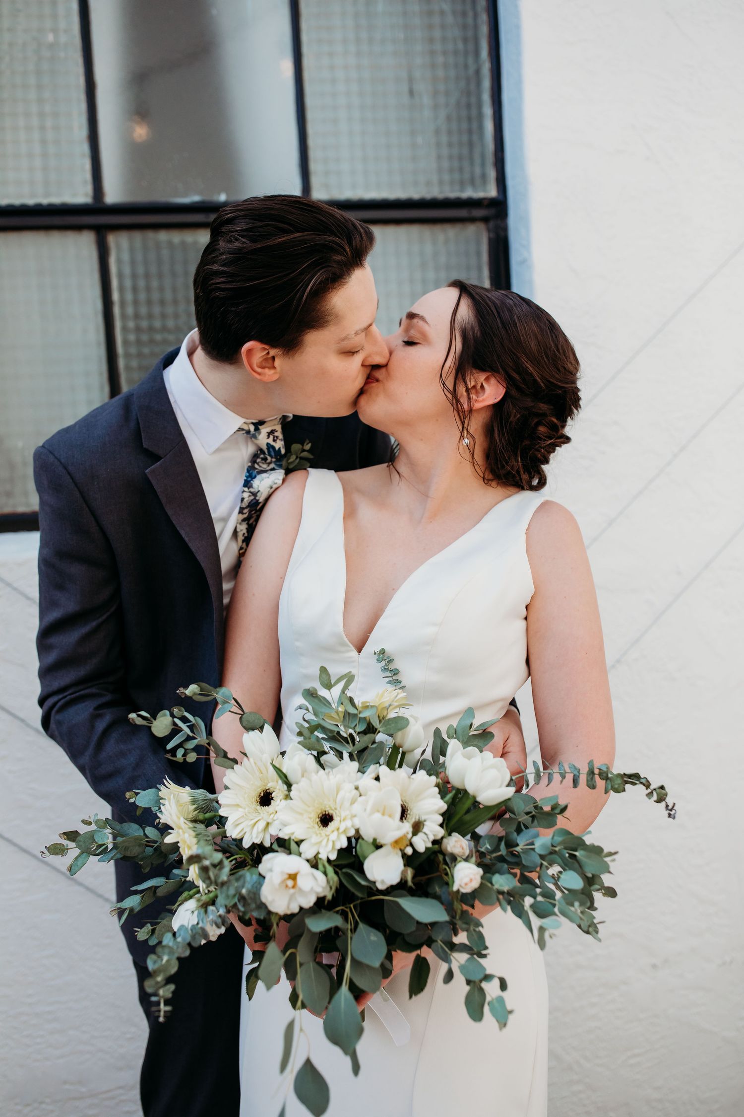A romantic wedding couple sharing a kiss while holding a beautiful white and green floral bouquet.
