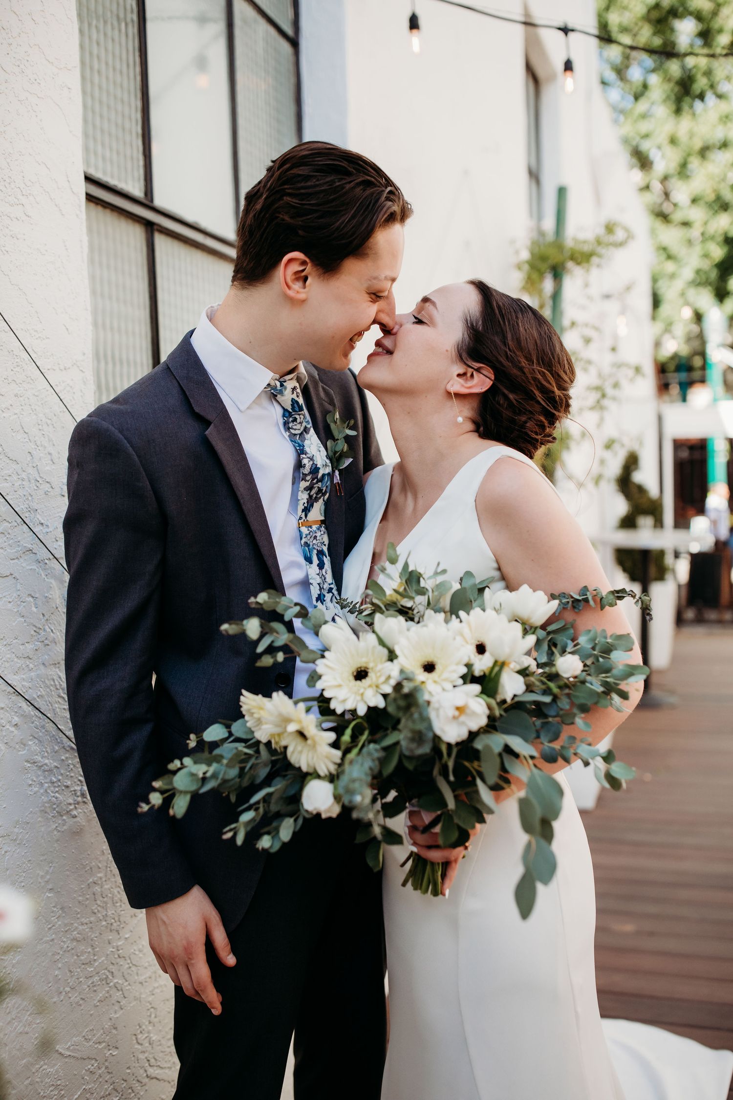 A bride and groom share a romantic kiss during their wedding photoshoot with eucalyptus and white floral bouquet accents.