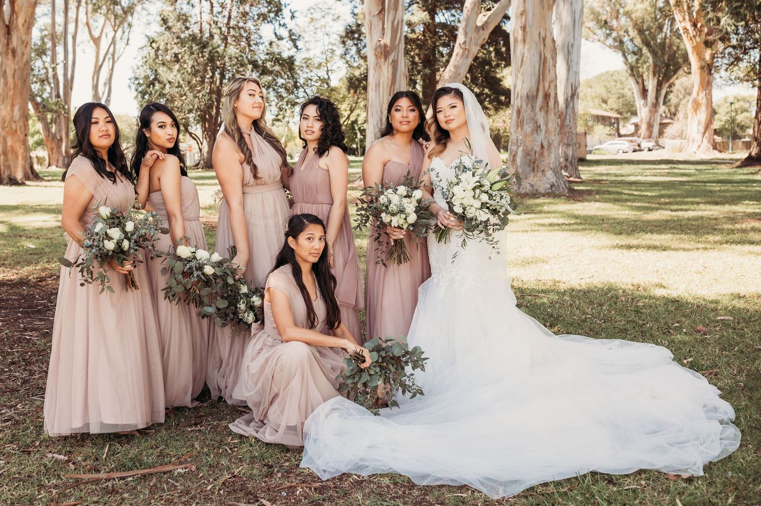 Wedding party in blush pink bridesmaid dresses pose with bride in white gown under eucalyptus trees with floral bouquets.