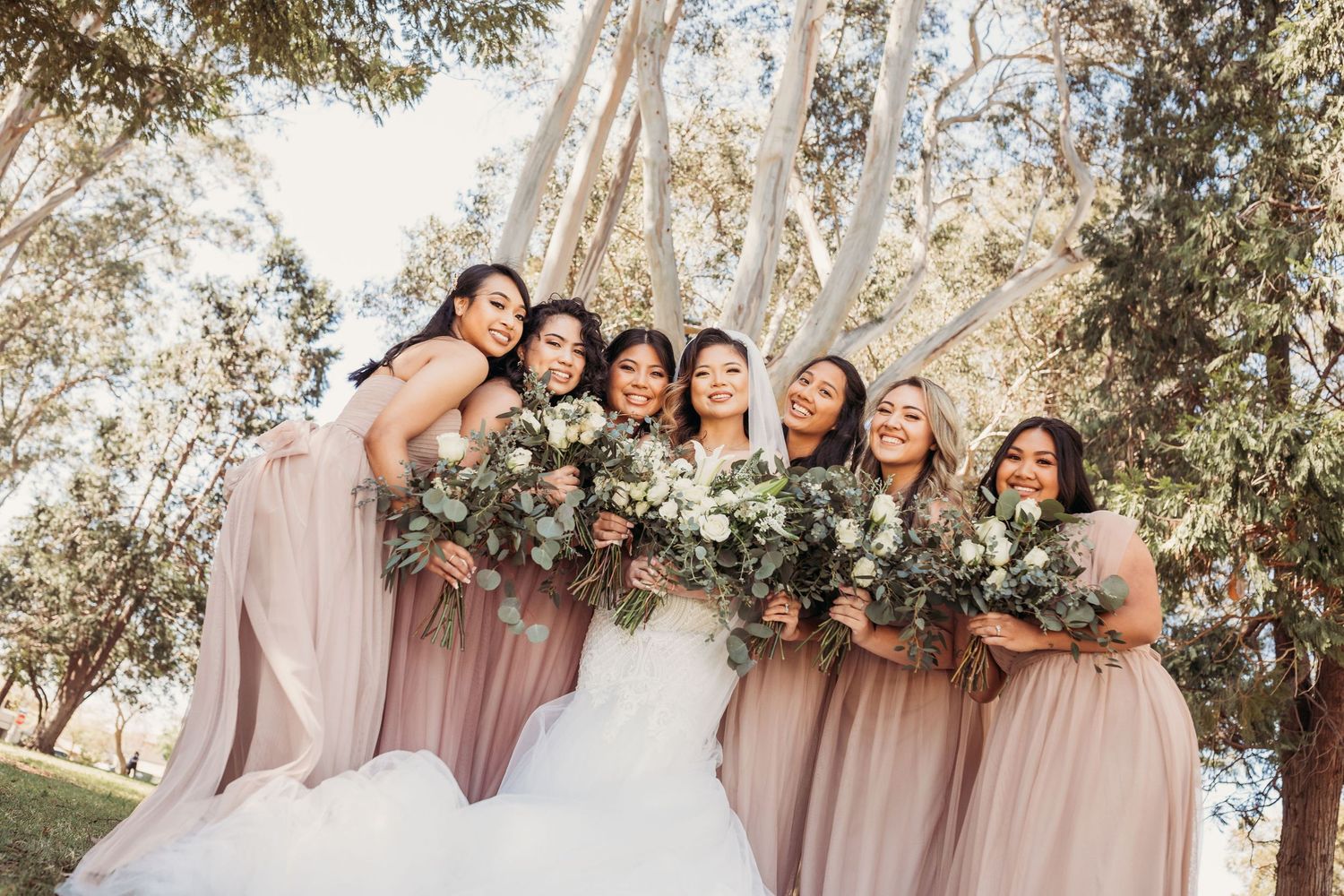 A wedding party in mauve bridesmaids dresses poses together outdoors beneath flowering trees in spring.