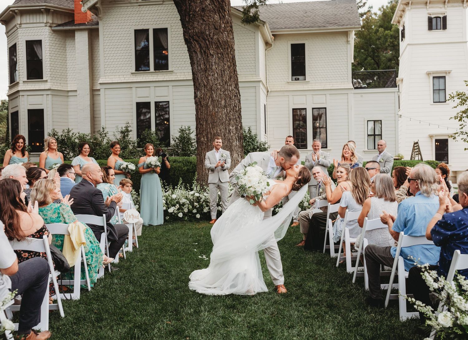 Guests celebrate during an outdoor wedding ceremony on a manicured lawn in front of a classic white house.
