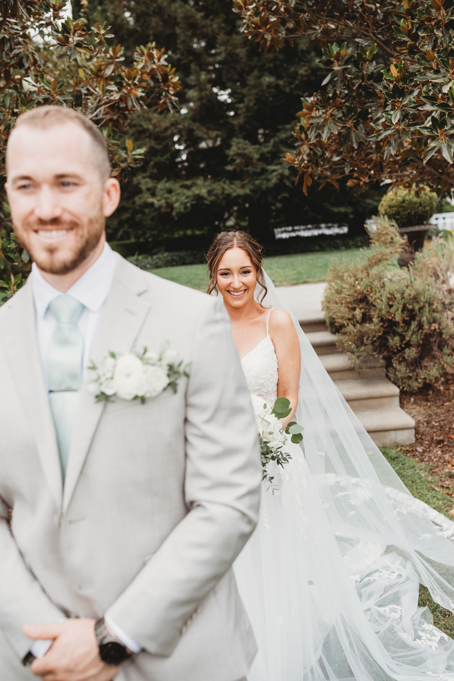 A bride and groom share a first look moment outdoors with her flowing white veil and his light gray suit with mint tie.