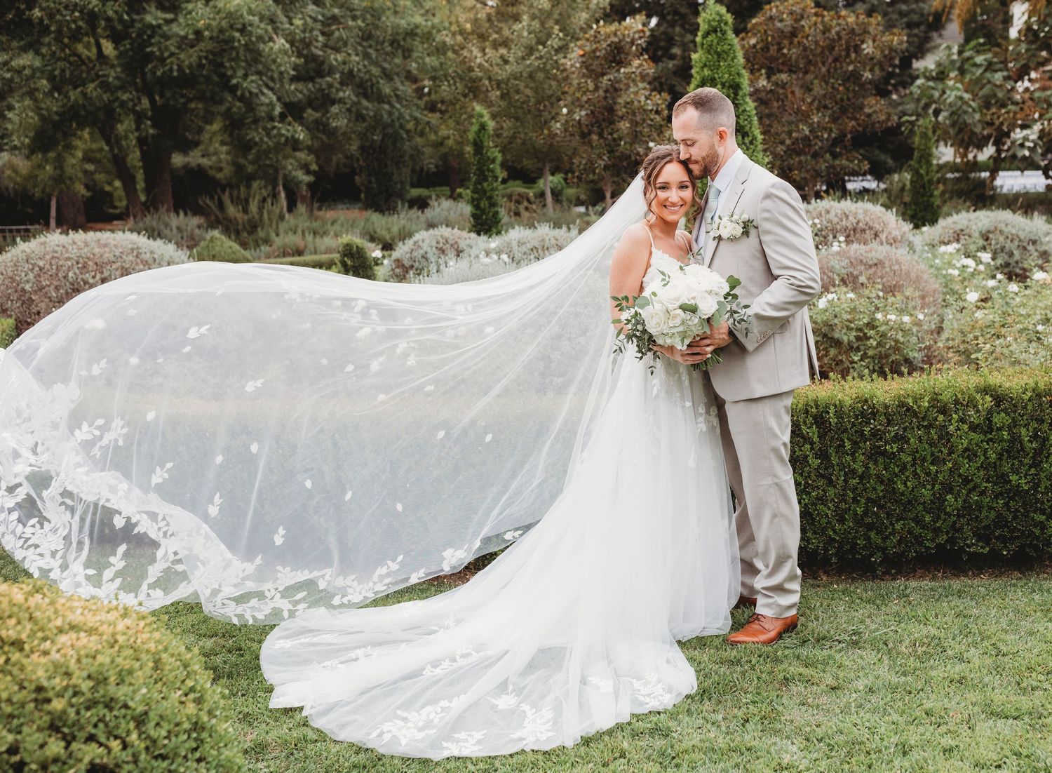 A couple shares an intimate moment in a garden setting with a flowing cathedral veil and bouquet of white flowers.