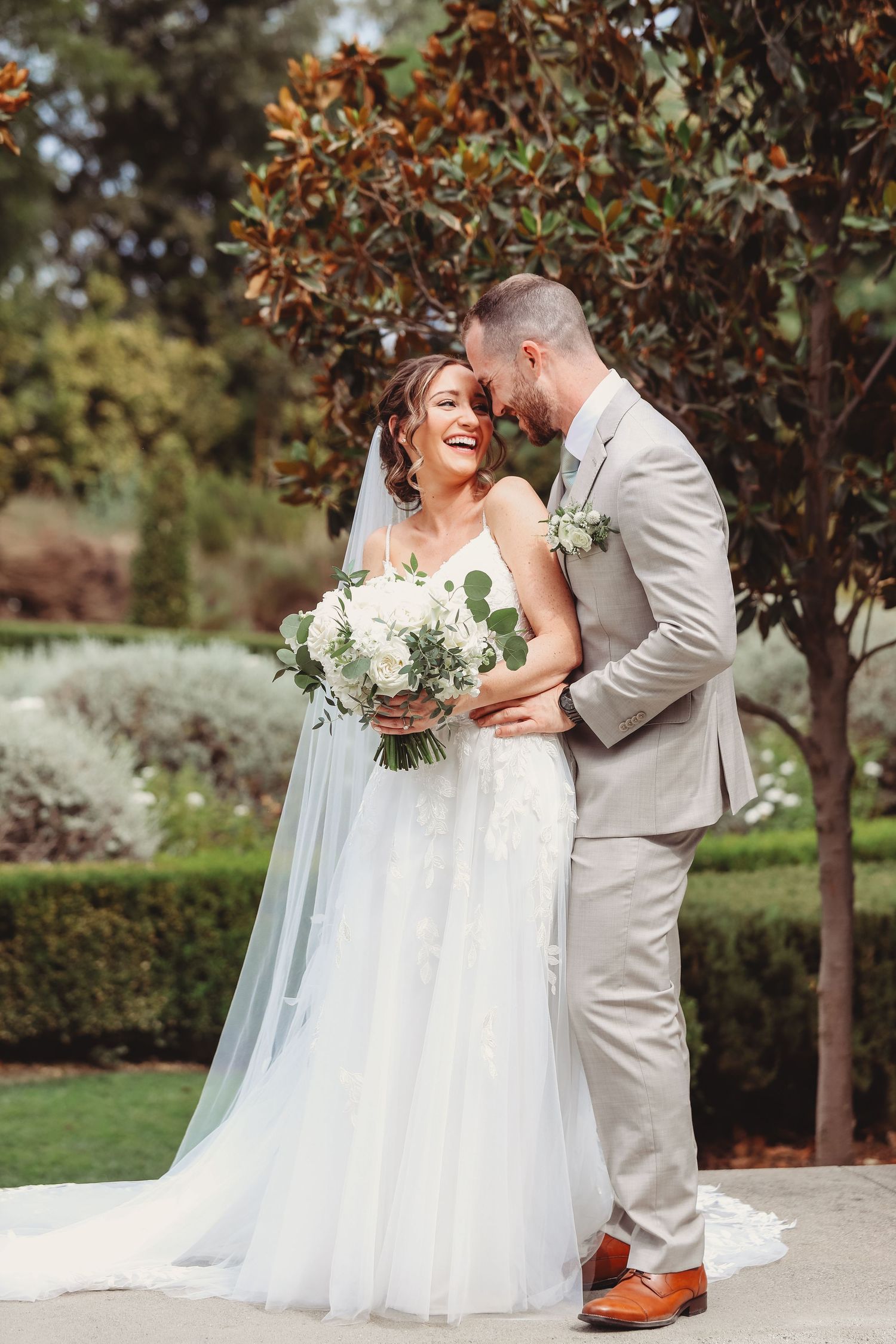 A joyful couple shares a candid laughing moment in their wedding attire on a garden walkway surrounded by trees.