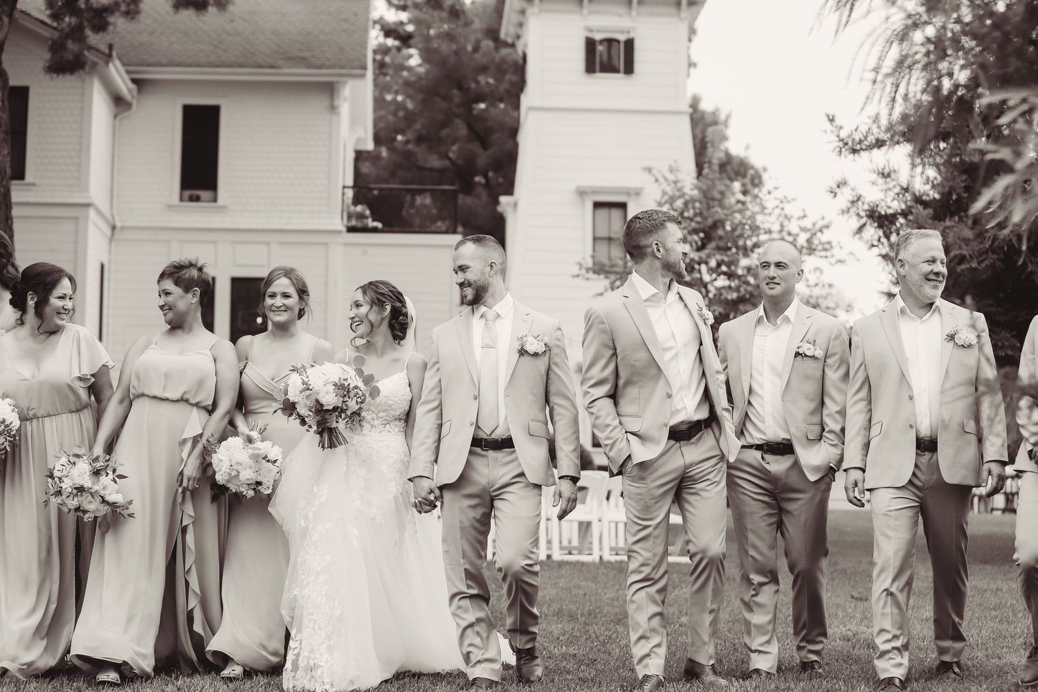 A wedding party in light colored attire poses outdoors in front of a white building in a vintage black and white photo.