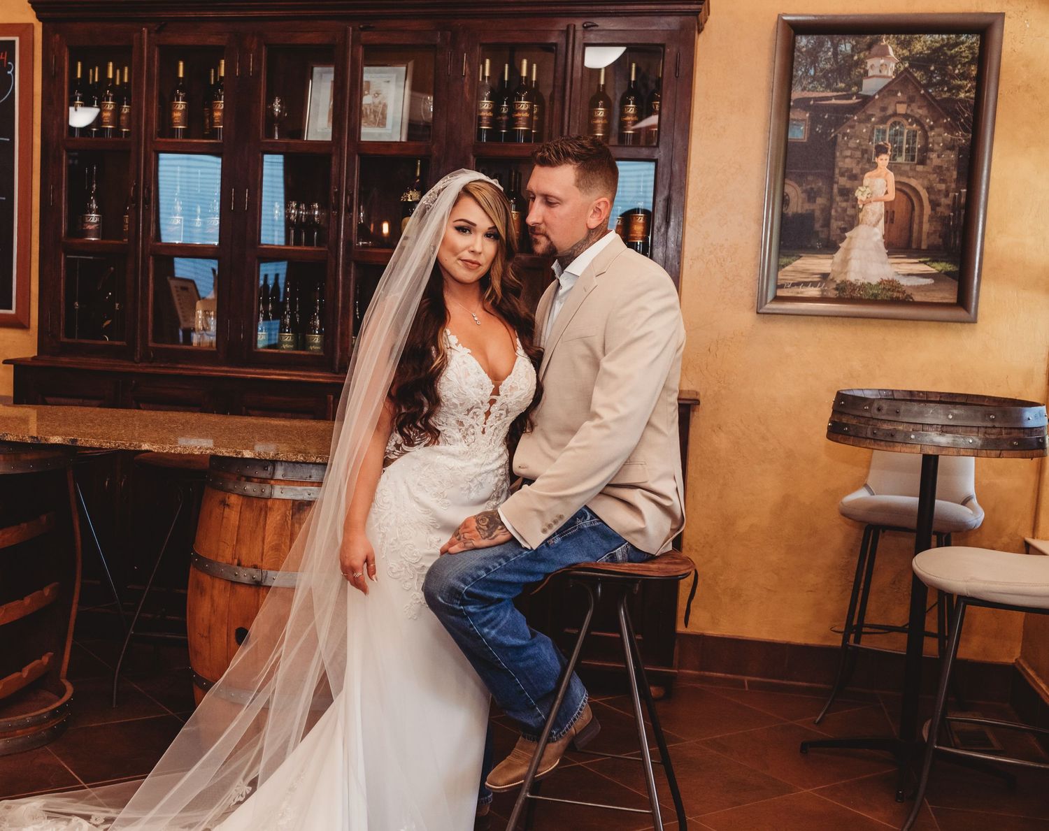 A couple in wedding attire sits together at a rustic bar with wooden decor and warm lighting in the background.