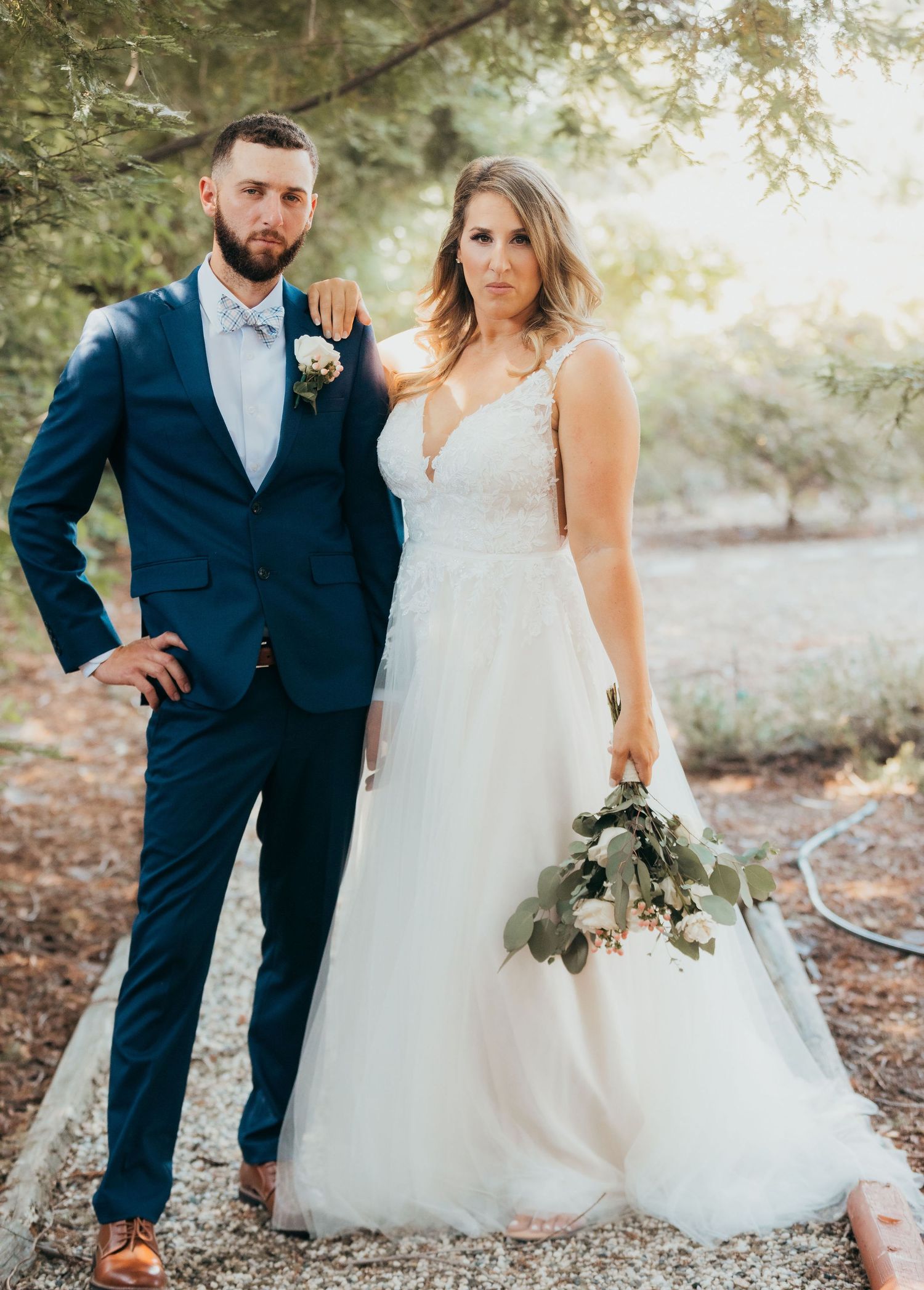 Couple in wedding attire posing outdoors at sunset with pine trees in background and rustic natural setting.