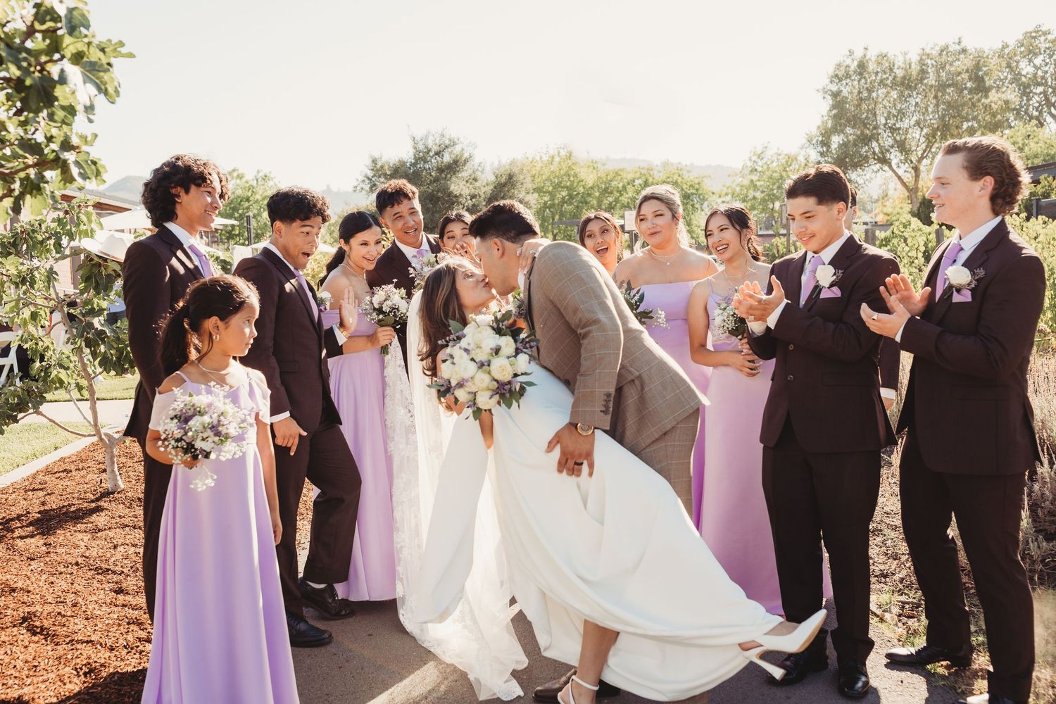 Wedding party in black tuxedos and lavender dresses pose together outdoors during sunset celebration.