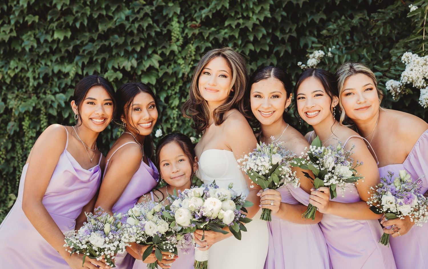 A wedding party in lavender bridesmaid dresses poses together with white floral bouquets in an outdoor garden setting.