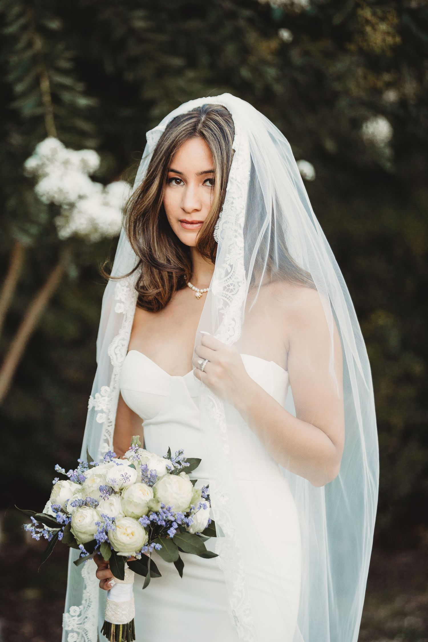 A bride in a white wedding dress holds a delicate bouquet of white peonies and blue flowers while wearing a flowing veil.