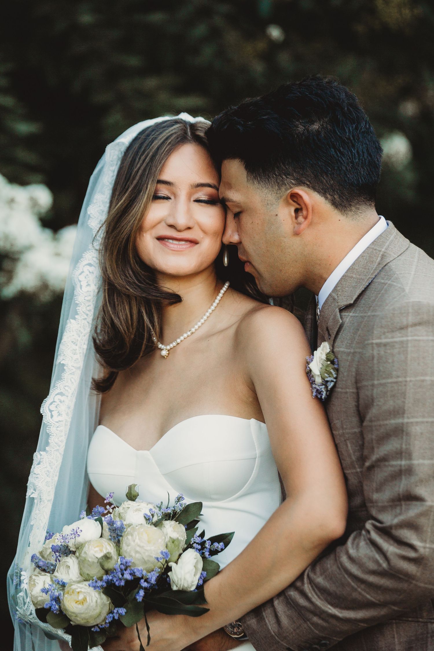 Newlyweds share intimate moment outdoors with bride in white dress holding blue and white bouquet.