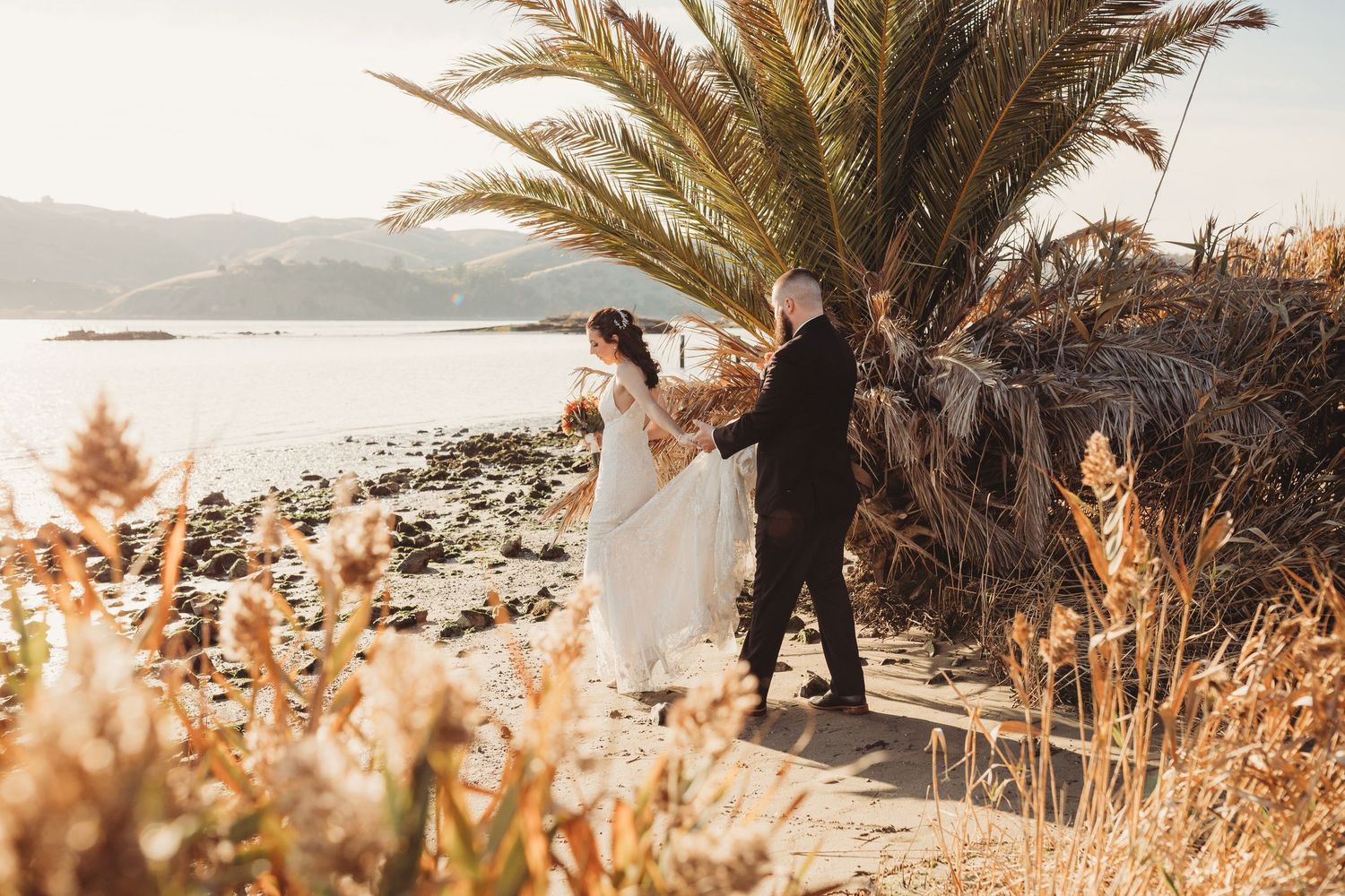A couple shares a romantic sunset moment on a rocky beach with a palm tree and golden grasses in the foreground.