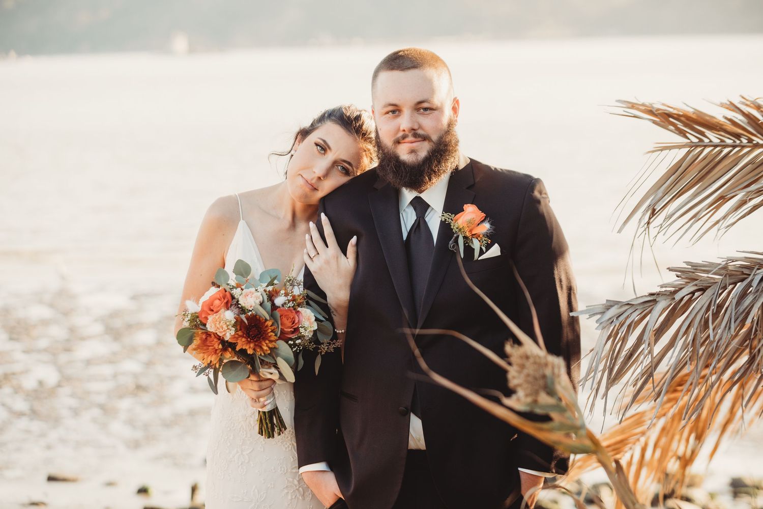 Newlyweds embrace on a sandy beach at sunset with palm fronds swaying in the background.