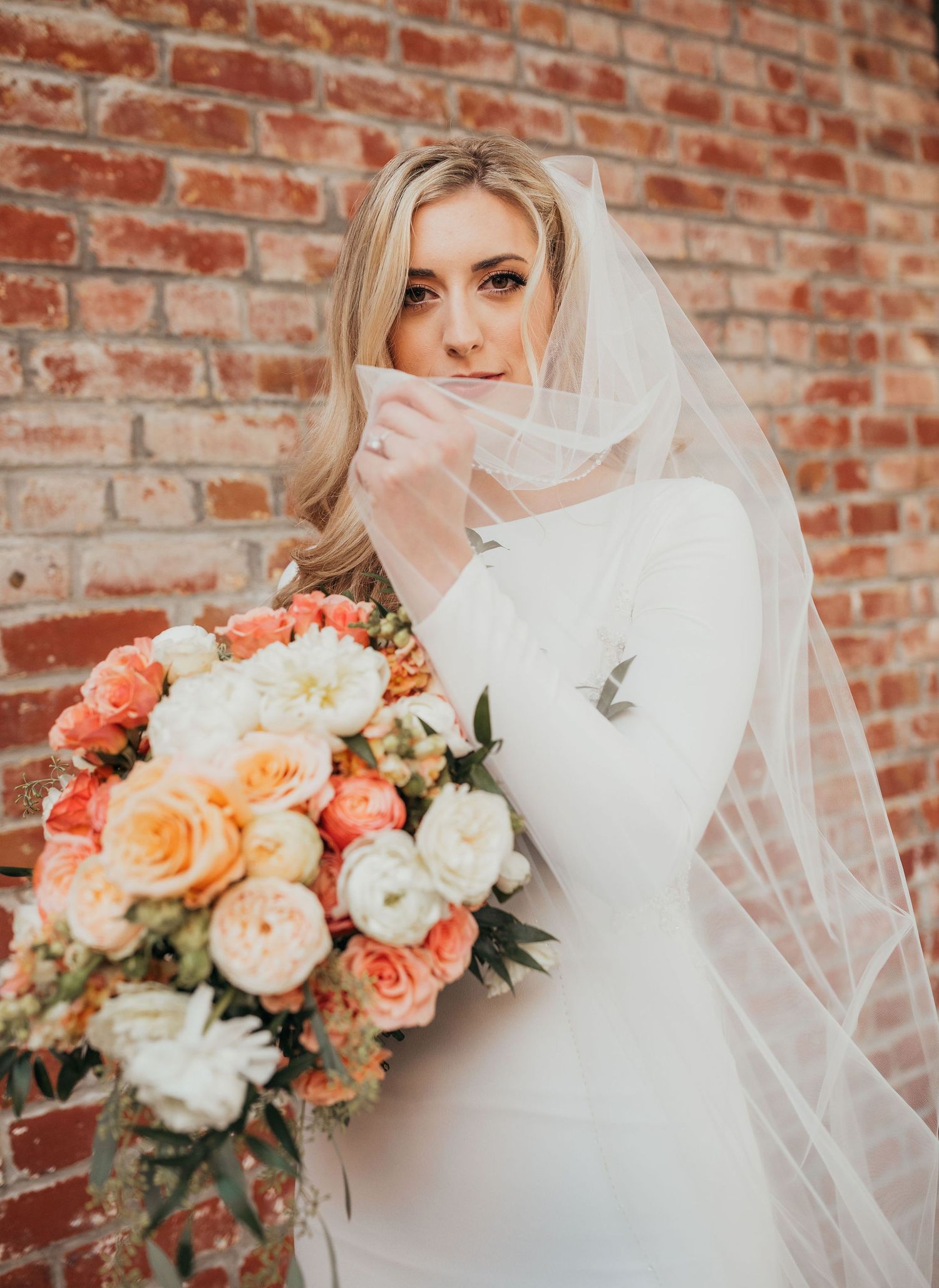 Romantic wedding portrait series against rustic brick wall with coral and peach rose bouquet and flowing white veil.