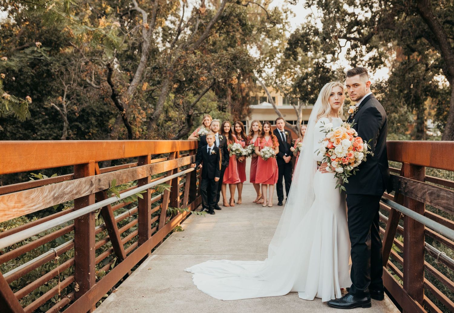 Wedding ceremony attendees gather on rustic wooden bridge during golden hour at outdoor forest venue.