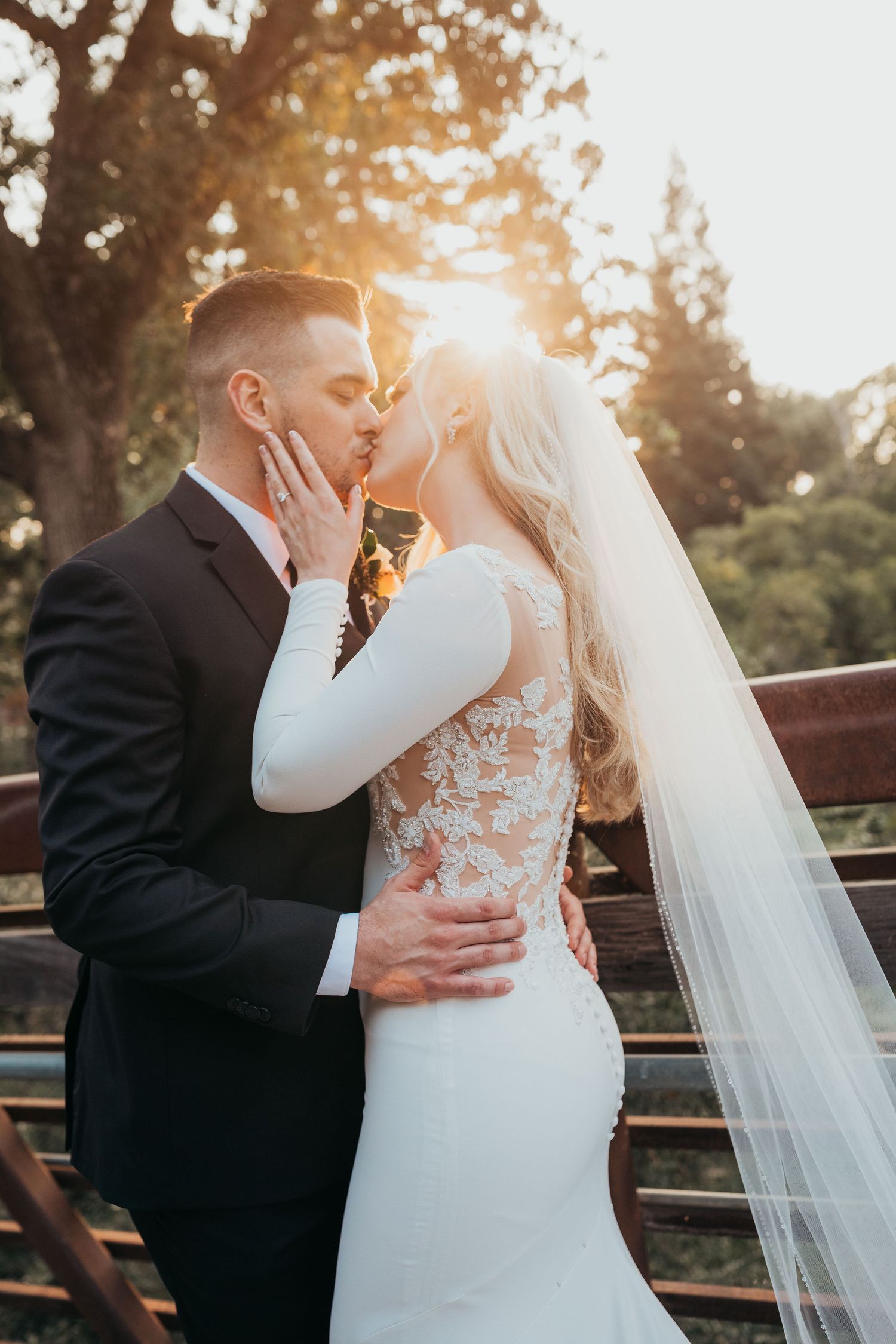 Romantic wedding couple sharing intimate kiss at sunset with flowing bridal veil and lace detail dress.